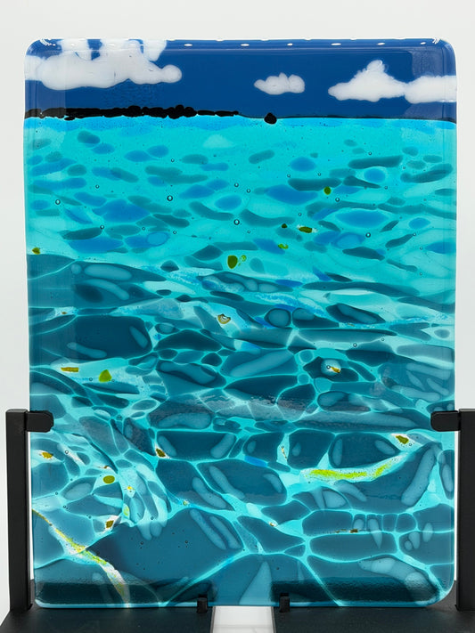 A square glass art piece depicting an underwater ocean scene with turquoise water, blue sky, white clouds, and small yellow fish, displayed on a black stand against a white background.