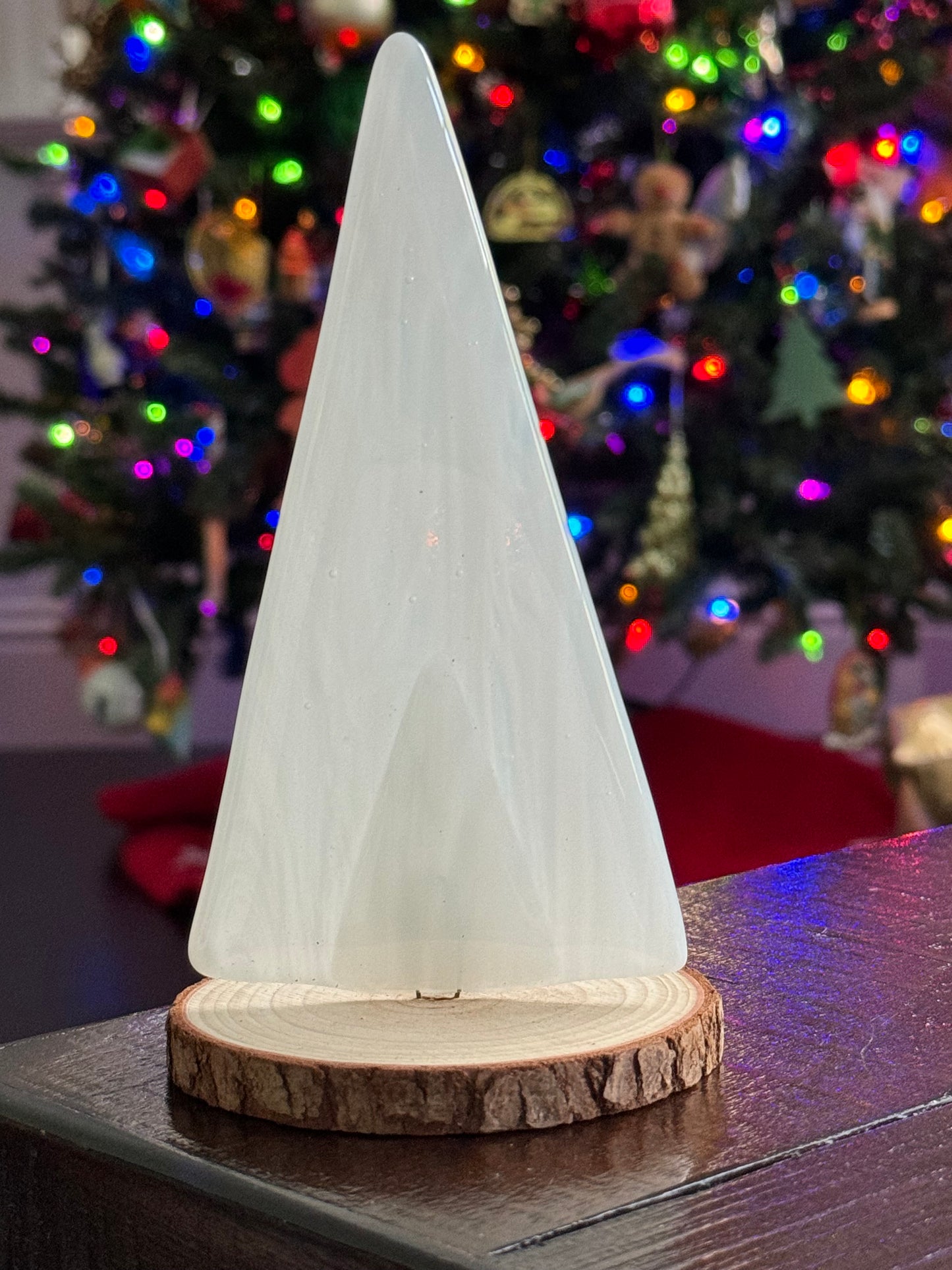 Small glass Christmas tree on a wood stand in front of a large lit Christmas tree