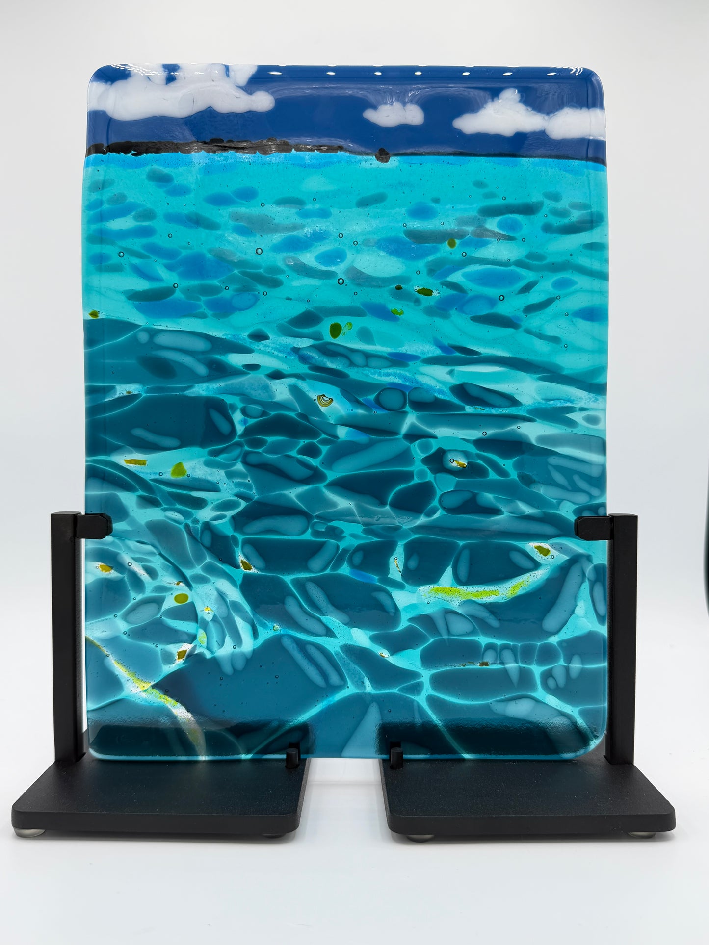 A rectangular glass art piece depicting a sunny blue ocean scene with ripples, light reflections, and white clouds on the horizon, displayed on a black metal stand against a white background.