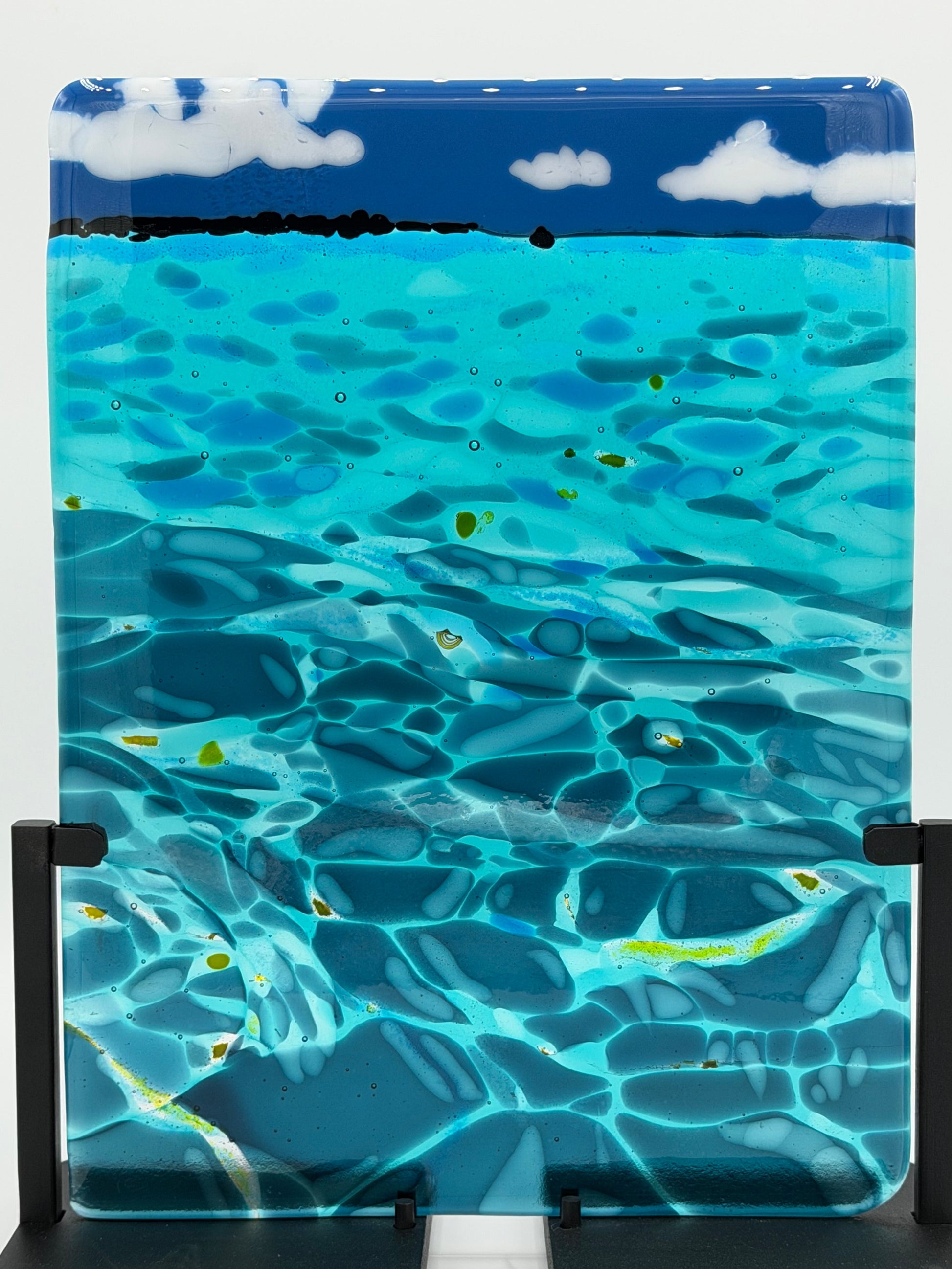 A square glass art piece depicting an underwater ocean scene with turquoise water, blue sky, white clouds, and small yellow fish, displayed on a black stand against a white background.