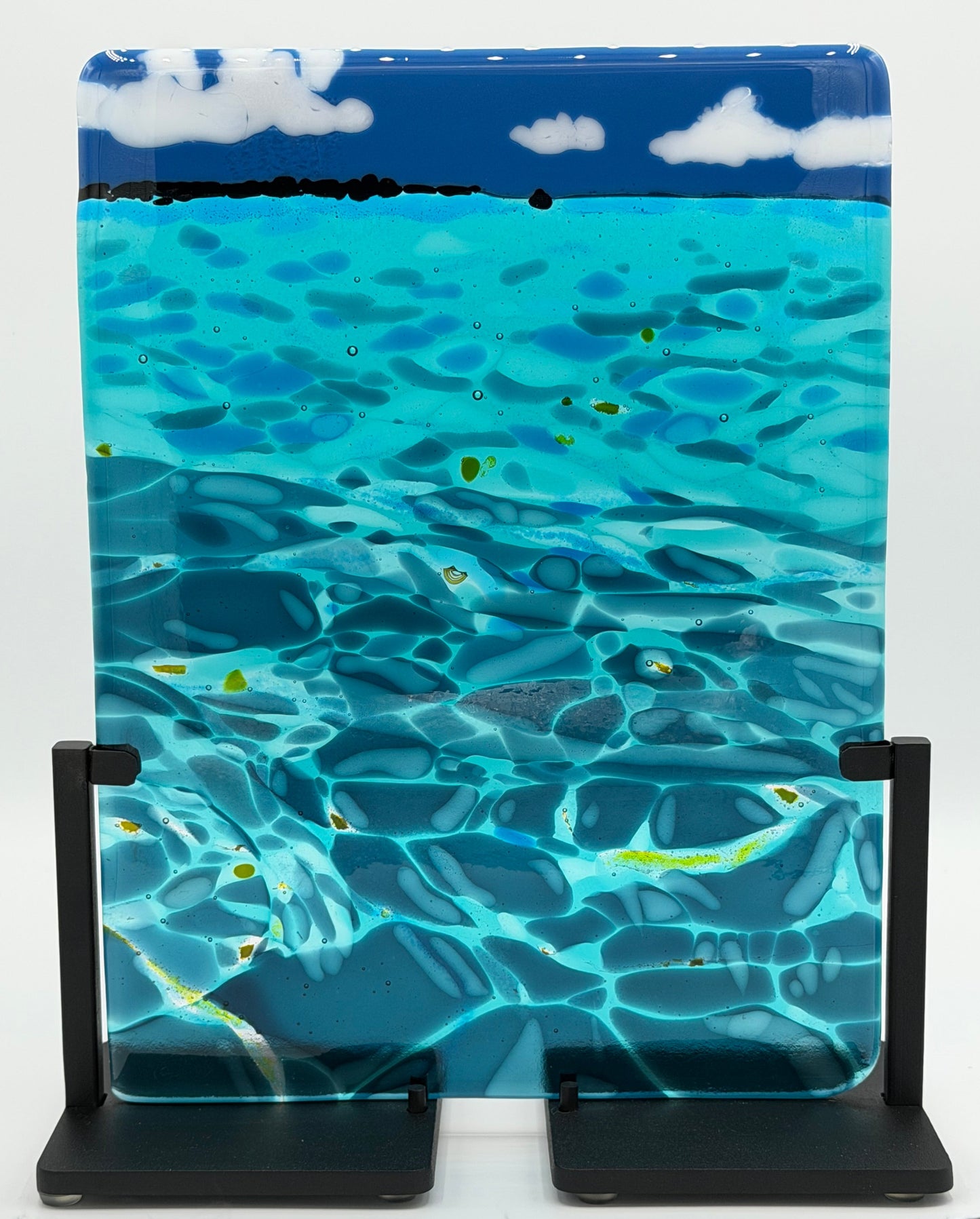 A rectangular glass art piece depicting a blue ocean scene with turquoise water, dark blue shadows, white clouds, and small yellow-green highlights, displayed on a black stand against a white studio background.