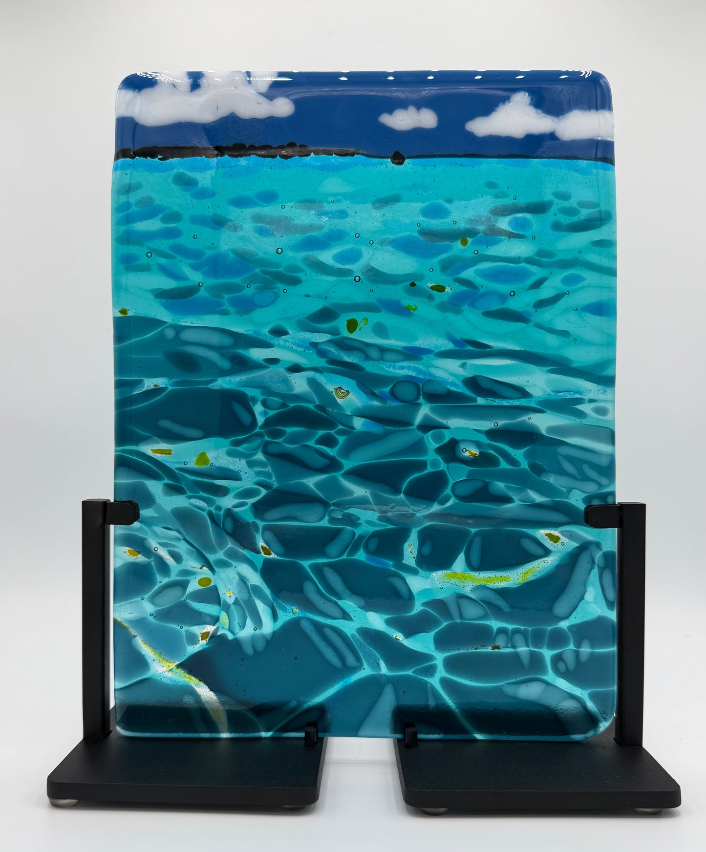 A rectangular glass art piece depicting a scenic ocean view with turquoise water, ripples, a dark blue horizon, and white clouds, displayed on a black metal stand against a white background.