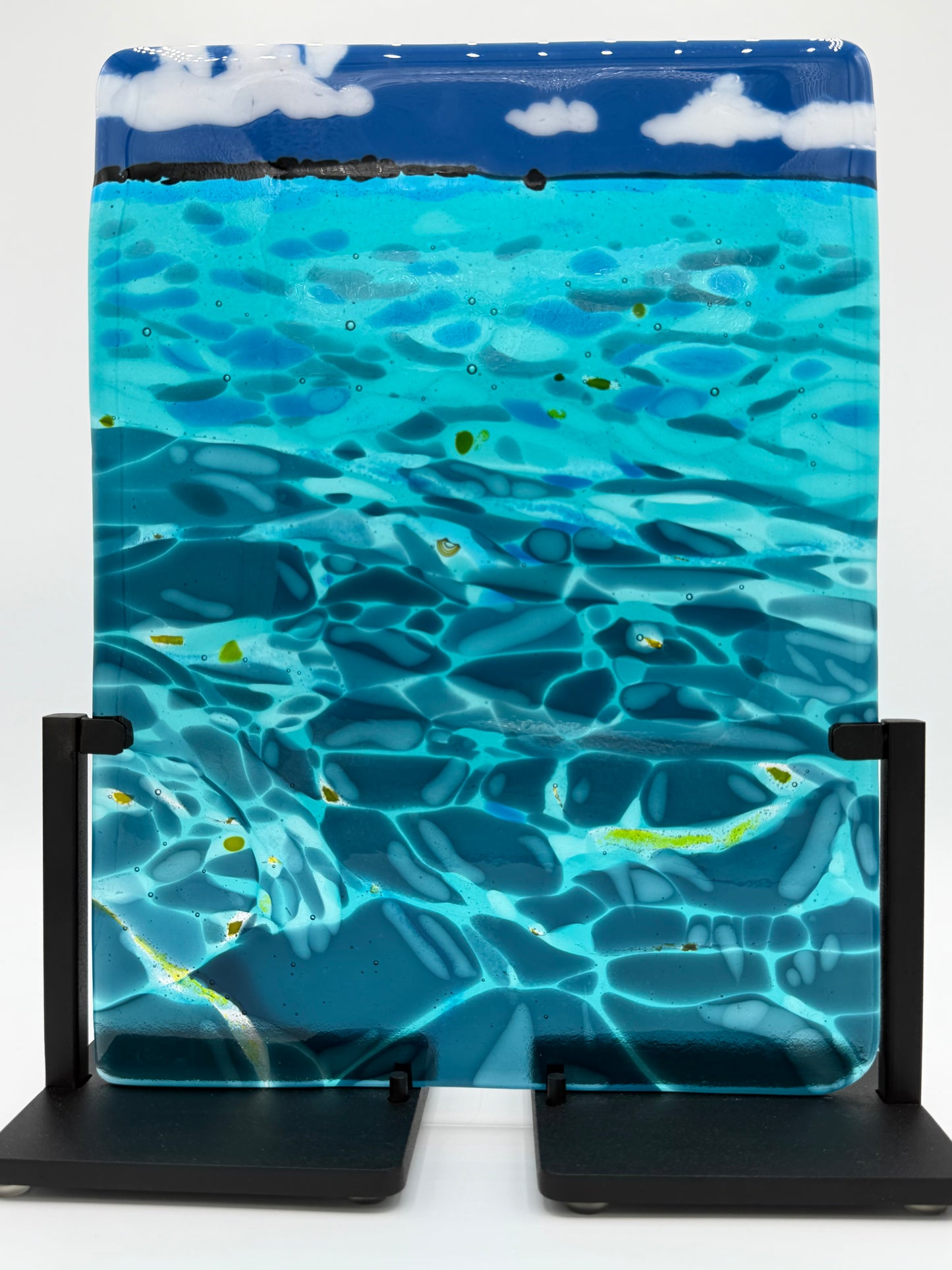 A rectangular glass art piece depicting a swimming pool scene with blue water, ripples, and white clouds at the top, displayed on a black stand against a white background.