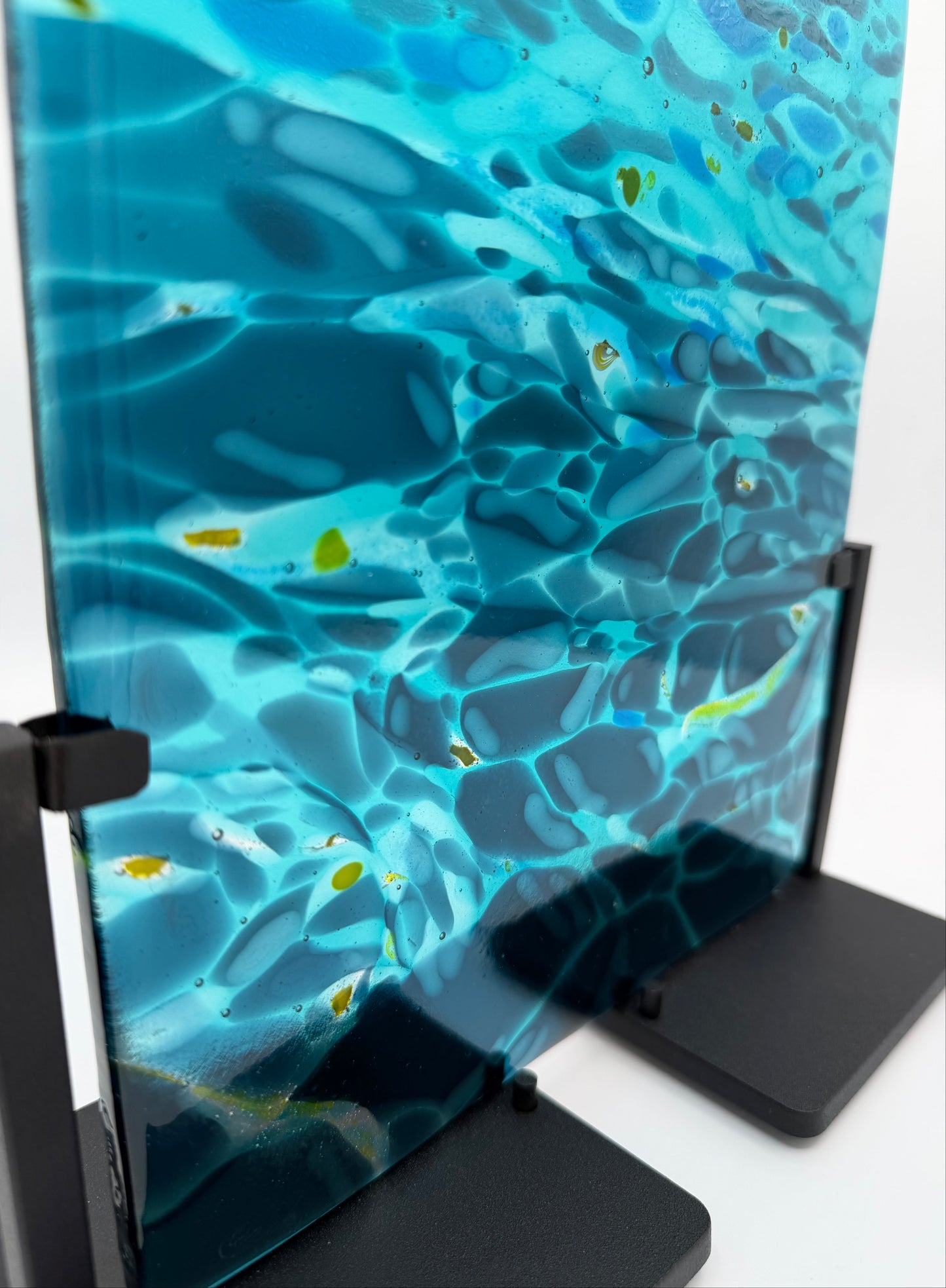 A vertical glass art panel with an abstract underwater wave pattern in shades of blue, teal, and black, mounted on a black metal display stand against a white background.