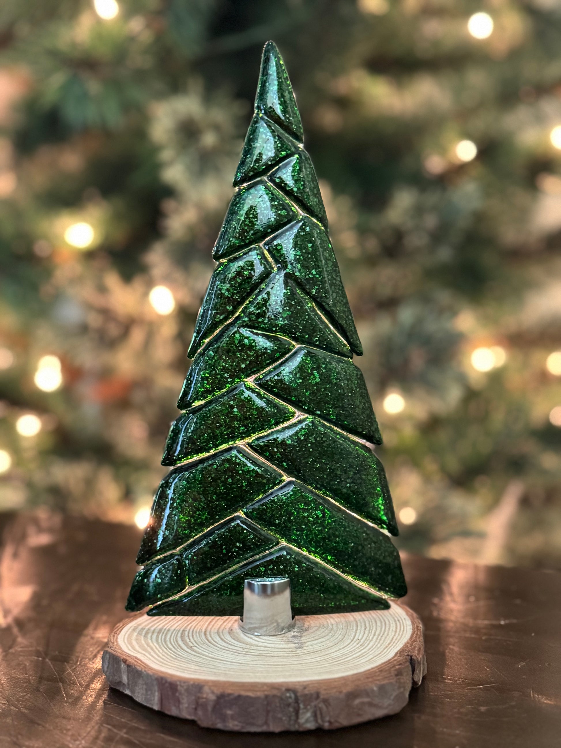 Green glass Christmas tree on a wooden base with blurred Christmas tree lights in the background