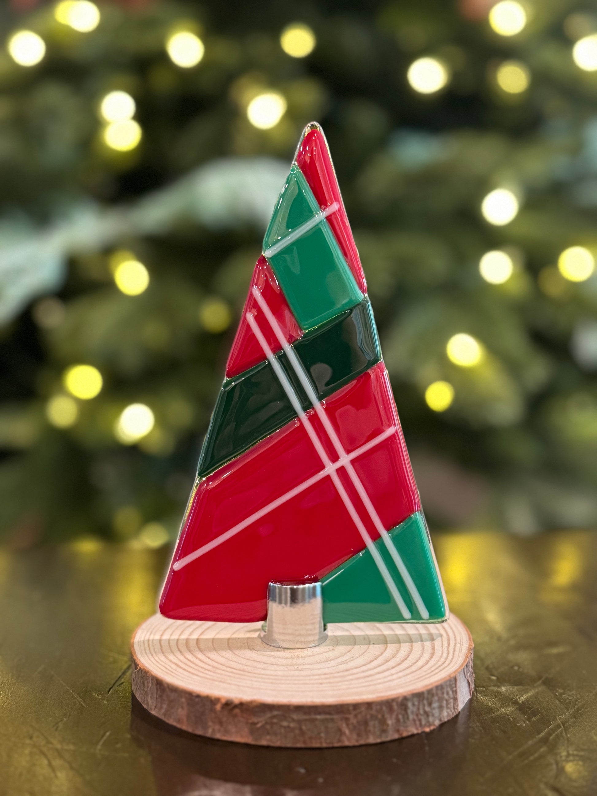 Small red and green plaid glass tree on a wooden base in front of a lit Christmas tree.