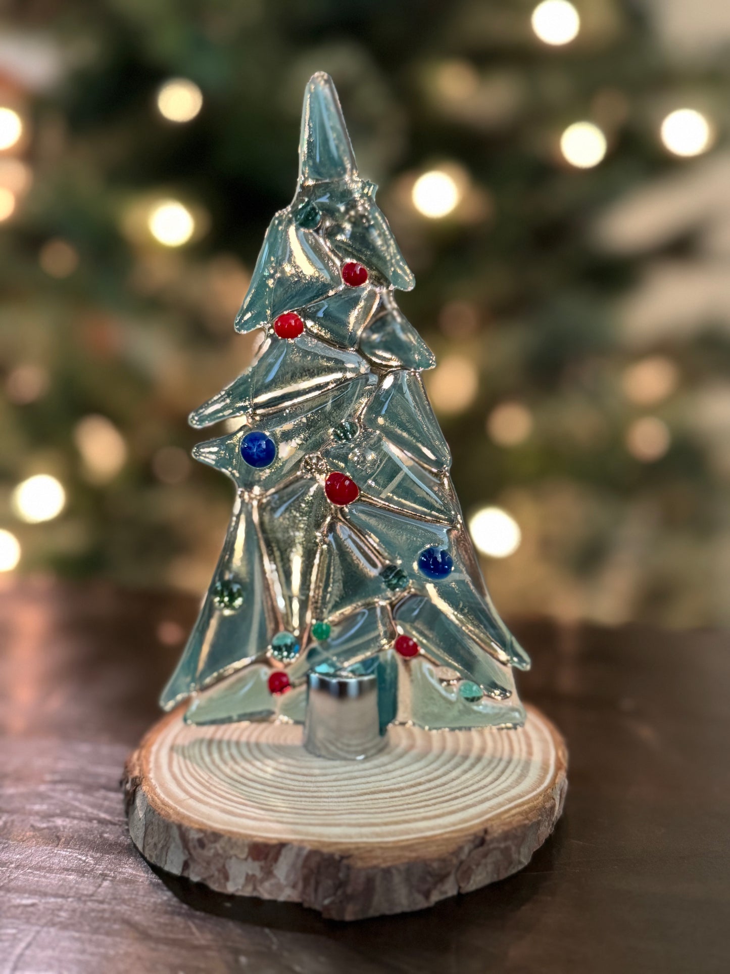 Sea green glass Christmas tree held on an unfinished wood stand by a chrome fastener. Christmas tree in background.