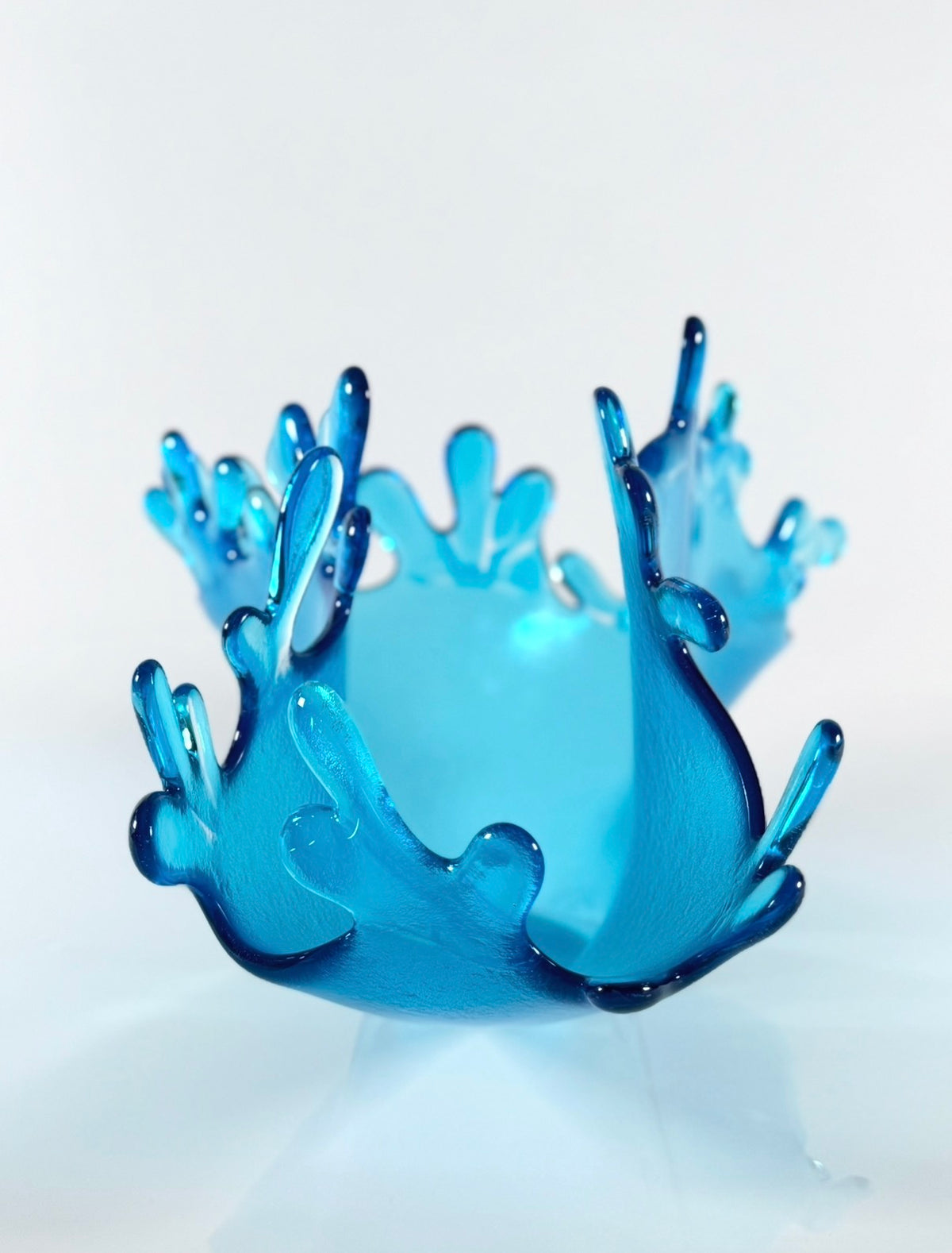 Sky blue glass sculpture depicting of a splash of water. Edge of glass resembles water droplets shown on white backdrop.