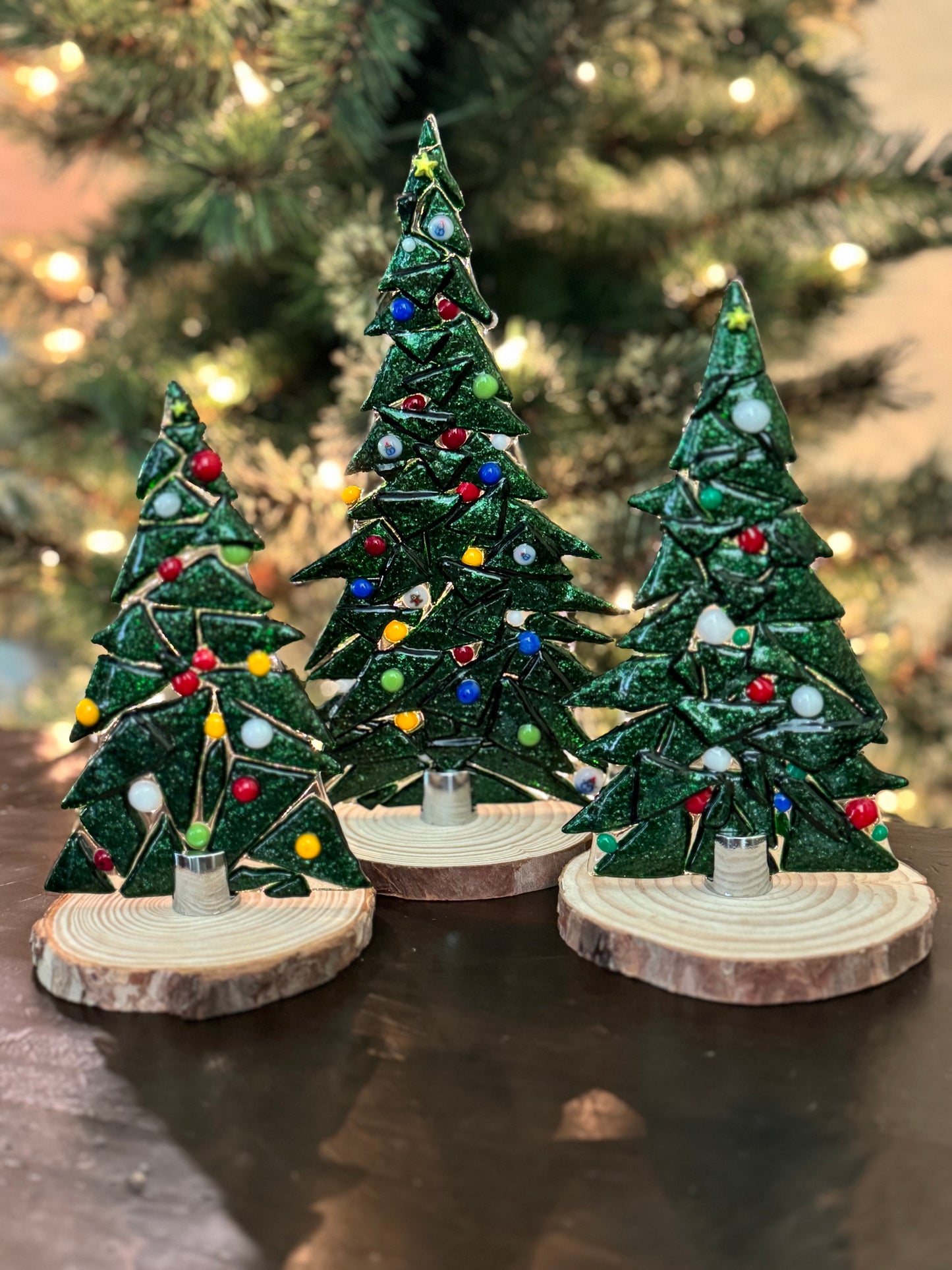 Three decorative Christmas trees with lights on wooden bases in front of a blurred Christmas tree background.